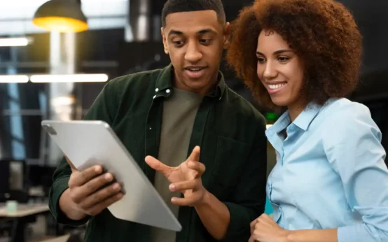 Imagem mostrando duas pessoas interagindo e adotando novas tecnologias através de um tablet em um ambiente moderno de trabalho.
