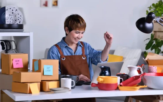 Imagem que ilustra como transformar um pequeno negócio em uma marca forte, com uma empreendedora sorrindo e interagindo com um computador, cercada por utensílios e caixas de embalagem.