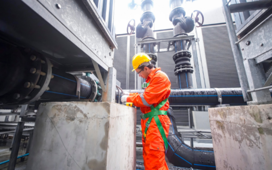 Trabalhador com capacete amarelo e uniforme laranja utiliza equipamentos de proteção enquanto realiza inspeção em tubulações industriais.
