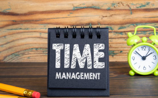 Bloco de notas com a frase “Time Management” ao lado de relógio verde sobre mesa de madeira.