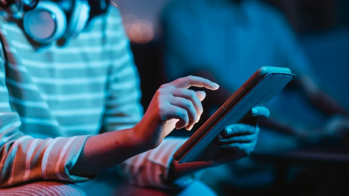 Mãos utilizando um tablet em um ambiente com iluminação azul, sugerindo Principais Tendências De Tecnologia Que Devem Ganhar Espaço Nos Próximos Anos e uso de dispositivos digitais